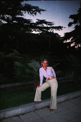 A young woman stands outdoors, wearing a light-colored button-down shirt tied at the waist, wide-leg trousers, and sandals. A...