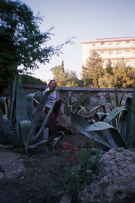 A woman in wide-leg trousers stands near a large agave plant in a stone-walled garden. A multi-story concrete building is vis...