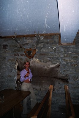 A woman in 1970s attire (bell-bottoms, button-down shirt) stands in a room with stone walls and a mounted stag head. Visible ...