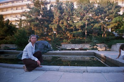 A woman in casual clothing kneels by a tiered fountain featuring a bronze sculpture, likely in a coastal resort setting. Arch...