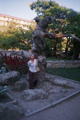 A woman poses beside a gnarled, sculptural tree set within a stone-edged garden. A building with red accents is visible beyon...
