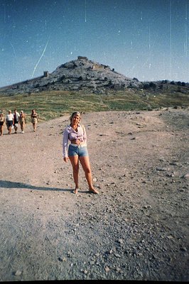 A young woman in denim shorts and a floral blouse poses on a gravelly slope, facing a rocky hilltop. A small group of people ...