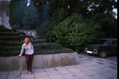 A woman stands barefoot on a paved area next to a low, concrete retaining wall densely planted with small flowers. A dark gre...