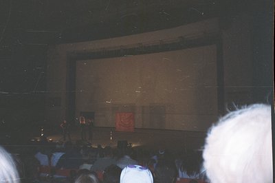 A blurry, low-light photograph depicts an audience inside a theater, viewed from a seat. A large cinema screen dominates the ...