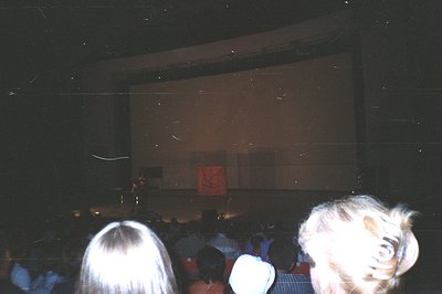 Low-light, candid shot of a theater audience. Viewers seated in red chairs face a large screen displaying a logo or emblem. A...