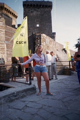 A woman in denim shorts and a cropped shirt poses before a stone tower with yellow flags. The architectural style suggests a ...