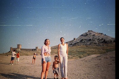 Two women and a young boy stand on a sandy beach with a rocky promontory topped by a fortress in the background. The image's ...