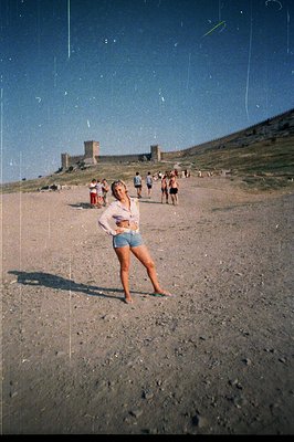 A young woman stands on a sandy beach, posing near a rocky hillside topped by a medieval castle ruin. Several figures are vis...