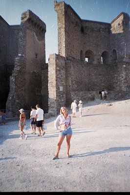 A young woman stands in a sandy courtyard before a weathered stone fortress with arched openings. Tourists explore the area. ...