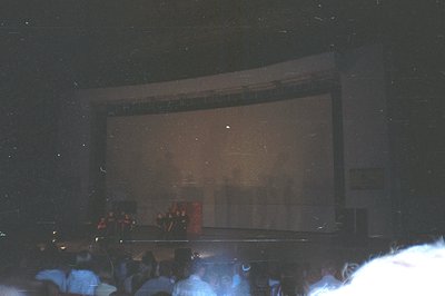 A darkened cinema interior; the screen dominates, showing a blurred image. Audience members are visible as indistinct forms i...