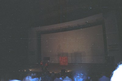 A dimly-lit movie theater scene. A large screen dominates the view, showing a red square shape, likely an intermission card o...