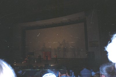 Concert stage view, showing three silhouetted figures performing against a backdrop of diffused lighting. Audience visible in...