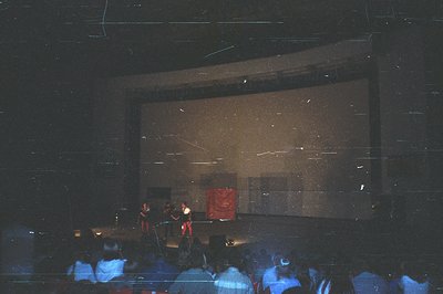 A grainy, low-light photograph depicts a stage performance within a darkened theater. Visible on screen are two figures in re...