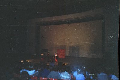 A performance stage is visible on a darkened theater screen, figures silhouetted against a patterned backdrop. Audience membe...
