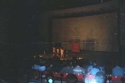 Performance stage with three figures in orange costumes, illuminated against a screen with a red graphic. Audience seated in ...