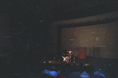 A performance stage is dimly lit, featuring a central figure in a red garment and a person in white standing next to them. Se...