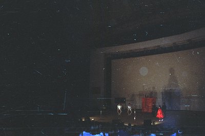 Performance stage, spotlight illuminates three figures in modern theatrical costumes. A backdrop features a large circular de...