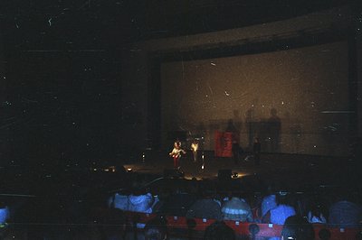 A stage performance captured from an audience perspective. Silhouetted performers are visible against a warm-toned backdrop, ...