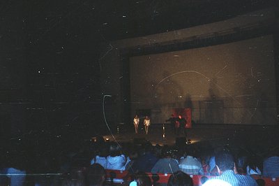 A theatrical performance or presentation is underway in a large, darkened auditorium. Three figures stand on a raised stage, ...