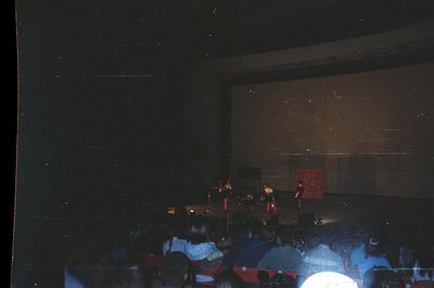 A stage performance with three figures in red attire is visible on a dimly lit platform. Audience members fill red-upholstere...