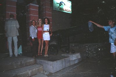 Three people pose in front of a neon "Baron" sign, likely a club or restaurant. Two women wear short, fitted dresses. A man i...