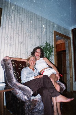 Two young women pose casually indoors, one seated in a large, patterned armchair, the other reclining atop. Distinctive 1970s...