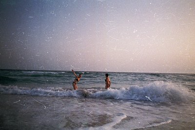 Two figures wade through breaking waves on a sandy shore. One raises an arm in exuberance; the other faces the ocean. Film gr...