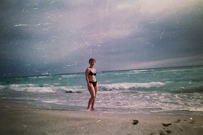 A young woman stands on a sandy beach facing the ocean. She's wearing a black bikini and appears to be wading in the shallow ...