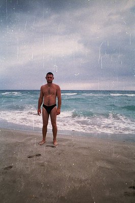 A man stands on a sandy beach, facing the camera. He's shirtless and wearing dark swim trunks. Waves gently roll onto the sho...