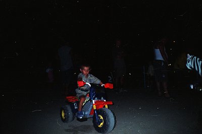 A young boy rides a red, blue, and black trike at night. Visible are onlookers in the background, blurred from motion or dist...