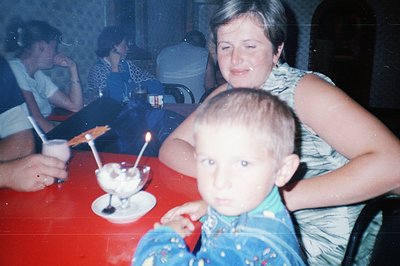 A young boy, likely in a patterned shirt, sits perched on a woman's lap at a table laden with a large dessert. The red tablec...