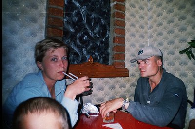 A candid moment at a table setting. A young woman with short, blonde hair lights a cigarette, while a man in a cap and polo s...