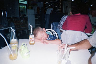 A young boy, wearing a blue overalls, sleeps with his head resting on a table. Two glass bottles of soda and a drink in a gla...