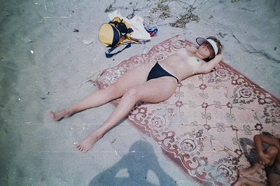 A woman in a black swimsuit and visor lounges on an ornate, patterned rug set on a beach. A small bag rests nearby. Likely a ...