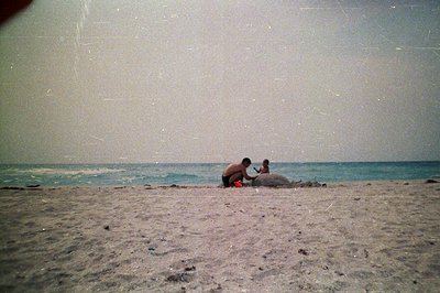 Two children build a sandcastle on a wide, sandy beach. A man stands nearby, partially visible. The scene is captured with a ...