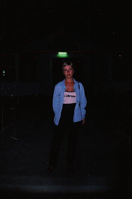 A young woman stands outside in near darkness, lit by a green glow above. She wears a Versace logo tank top, a light blue bla...