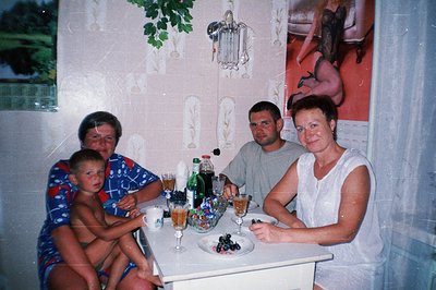 A family portrait: a young boy sits with his parents at a small, white table set with drinks & fruit. The interior features a...