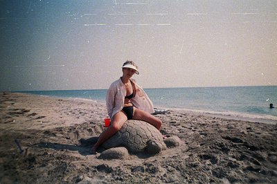A woman seated atop a sand sculpture resembling a sea turtle on a sandy beach, ocean backdrop. She wears a bikini, visor, and...