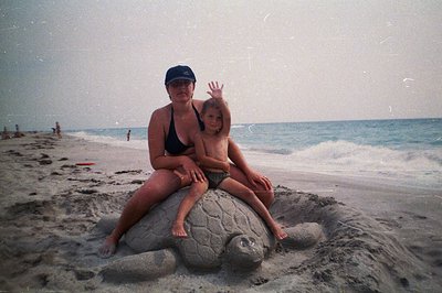 A woman and young boy sit atop a large sand sculpture of a sea turtle on a beach. Both are in swimwear; the woman wears a bas...
