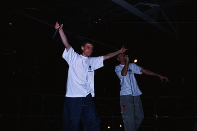 Two men perform on stage, raising arms and gesturing while rapping into microphones. They wear casual, baggy clothing – white...