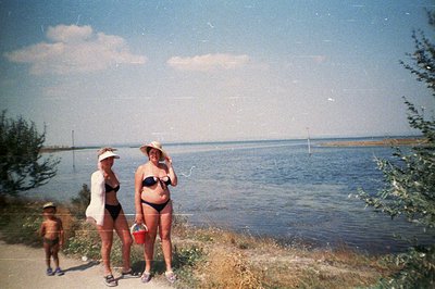 A lakeside scene captures two women in swimwear, standing alongside a young boy. The women wear 1970s-era swimwear and attire...