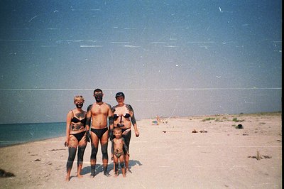 Family portrait on a sandy beach, covered in dark mud or clay. Three individuals—two adults and a young child—stand facing th...