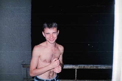 Young man, shirtless, stands with hands on hips; visible chest hair. Exterior setting with concrete wall and metal railing. L...