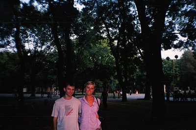 Two young adults stand in a large, leafy park, likely a resort area or seaside promenade. Mature trees frame the scene with b...