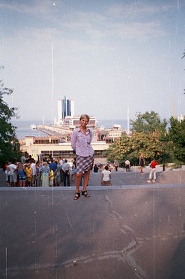 A young man stands on a tiered pathway, facing the camera. Behind him, a large seaside resort complex with distinctive modern...