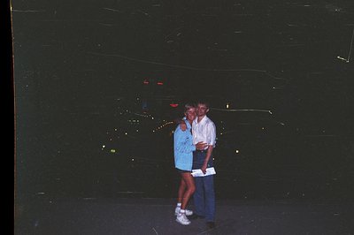A casually dressed couple stands close, embracing, in front of a brightly lit city backdrop, likely at night. The woman wears...