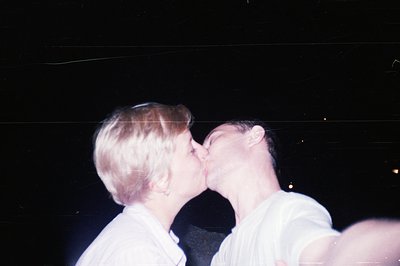 Close-up shot of a young couple kissing at night. Both are wearing simple white t-shirts. Grainy aesthetic suggests a snapsho...