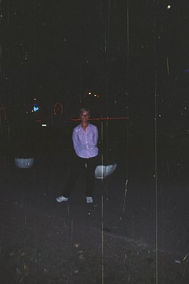 Man in a patterned button-down shirt & dark pants stands outdoors, likely in a city setting at night. Visible blurred neon li...
