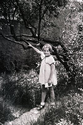 A young girl in a dotted dress and Mary Jane shoes stands on a stone path amidst lush foliage. She holds a small envelope or ...