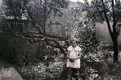 Young girl in a patterned dress stands amidst lush foliage, posed near a tree. The photograph, likely a candid portrait, reve...
